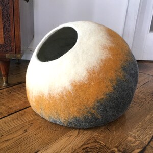 May include: A round, felted cat bed with a large opening. The bed is a gradient of white, yellow, and gray.