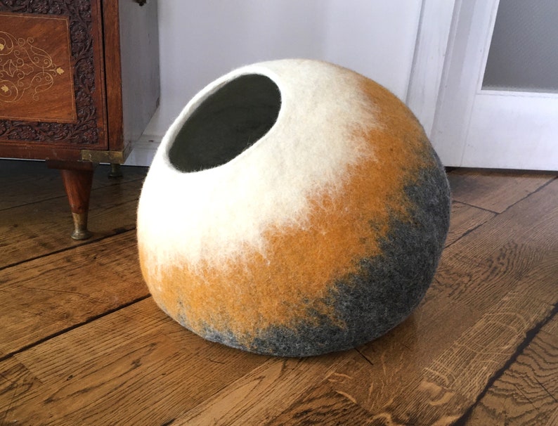 May include: A round, felted cat bed with a large opening. The bed is a gradient of white, yellow, and gray.