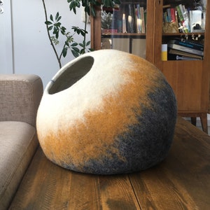May include: A round, felted cat bed with a white, yellow, and gray ombre design. The bed has a large opening for the cat to enter.