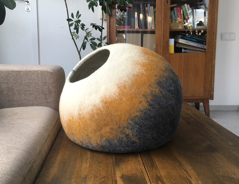 May include: A round, felted cat bed with a white, yellow, and gray ombre design. The bed has a large opening for the cat to enter.