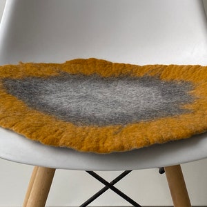 May include: A round, felted chair cushion with a gray center and a yellow outer edge. The cushion is sitting on a white chair.