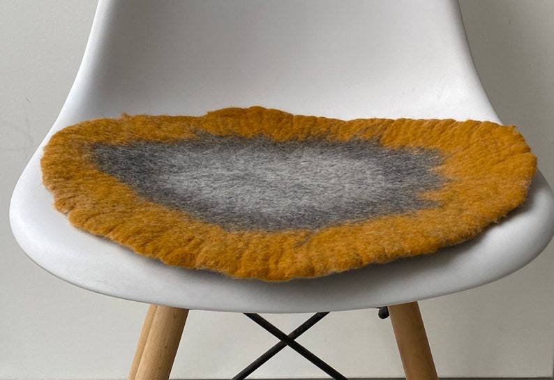 May include: A round, felted chair cushion with a gray center and a yellow outer edge. The cushion is sitting on a white chair.