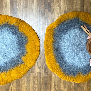 May include: Two round, felted wool placemats in shades of gray and yellow. The placemats are on a wooden surface. One placemat has a wooden bowl and chopsticks on top.