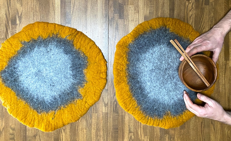 May include: Two round, felted wool placemats in shades of gray and yellow. The placemats are on a wooden surface. One placemat has a wooden bowl and chopsticks on top.