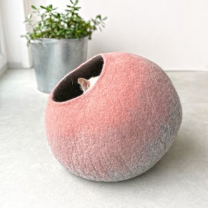 Peut inclure: Un lit pour chat en feutre rose et gris en forme de sphère. Un chat est partiellement visible à l'intérieur du lit.
