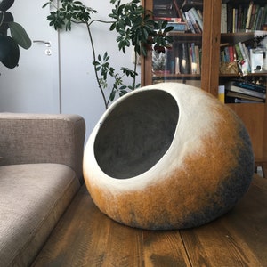 May include: A large, round, felted pet bed in shades of gray, white, and brown. The bed has a large opening for a pet to enter.