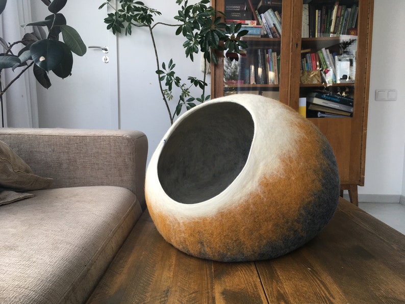 May include: A large, round, felted pet bed in shades of gray, white, and brown. The bed has a large opening for a pet to enter.
