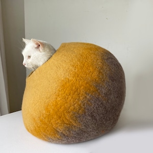 May include: A round, felted cat bed in shades of brown and yellow. The bed is made of wool and has a soft, fuzzy texture.