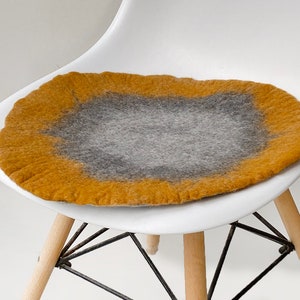 May include: A round, gray and mustard yellow felt chair cushion. The cushion is placed on a white chair with wooden legs.