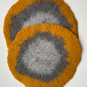 May include: Two round felt coasters in shades of gray and mustard yellow. The coasters have a circular design with a darker gray center and a lighter gray outer ring. The outer ring is surrounded by a band of mustard yellow.