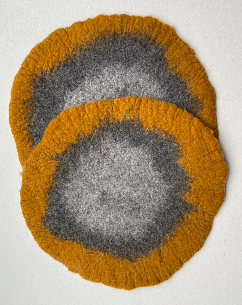 May include: Two round felt coasters in shades of gray and mustard yellow. The coasters have a circular design with a darker gray center and a lighter gray outer ring. The outer ring is surrounded by a band of mustard yellow.