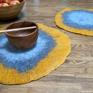 May include: Two round, felted placemats in shades of yellow and gray. The placemats have a unique, marbled design. A wooden bowl with a wooden spoon sits on one of the placemats.