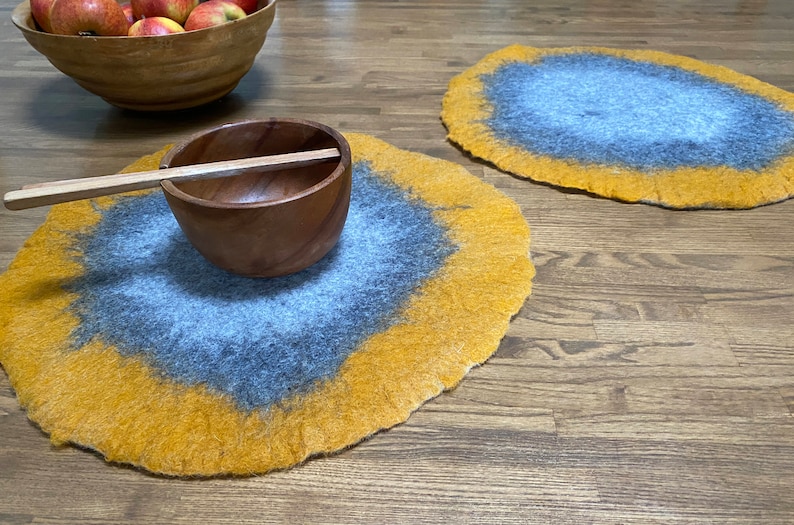 May include: Two round, felted placemats in shades of yellow and gray. The placemats have a unique, marbled design. A wooden bowl with a wooden spoon sits on one of the placemats.