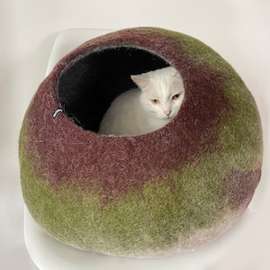 May include: A green and brown felted cat bed with a white cat resting inside. The bed is shaped like a dome and has a soft, fuzzy texture.