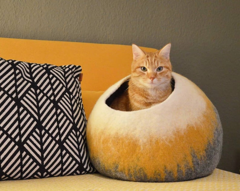 May include: A ginger cat sits inside a white, yellow, and gray felted cat bed. The bed is round and has a hole for the cat to enter. The cat is looking directly at the camera.