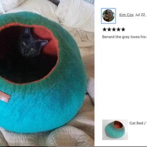 May include: A teal and orange cat bed made of felted wool. A gray cat is inside the bed. The bed is on a white and beige blanket. The text "Cat Bed / Cat Cave / Kitty Hou..." is visible in the image.