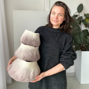 Puede incluir: Una mujer sostiene tres cuencos de fieltro apilados. Los cuencos son degradados, pasando de un beige claro a un marrón más oscuro. Los cuencos están hechos a mano y tienen una textura suave y peluda.