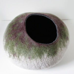 May include: A felted wool pet bed in shades of green, purple, and white. The bed has a round shape with a black felt lining inside the opening.