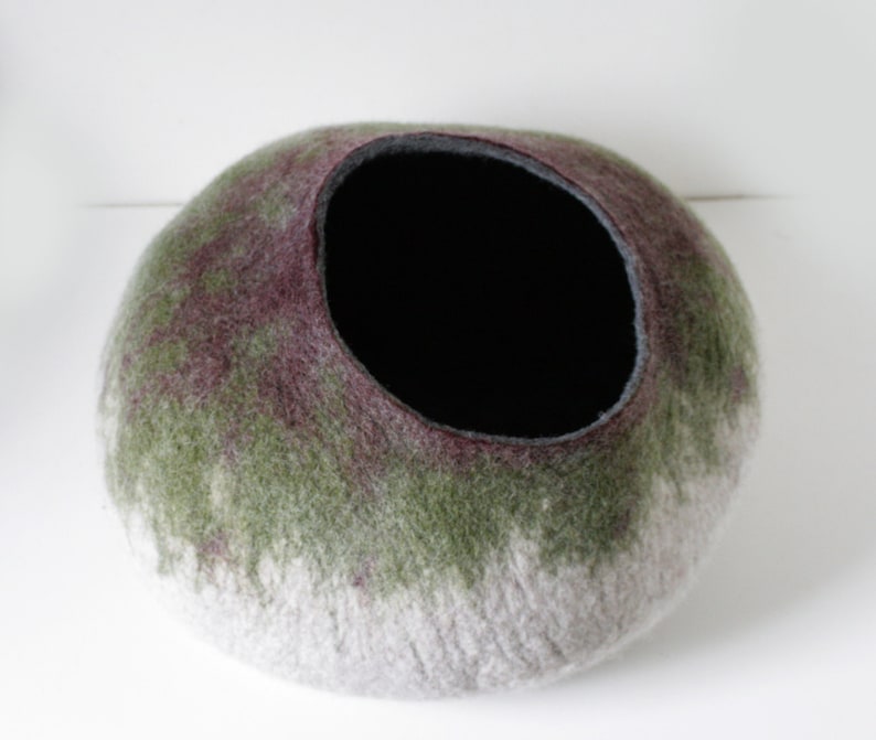 May include: A felted wool pet bed in shades of green, purple, and white. The bed has a round shape with a black felt lining inside the opening.