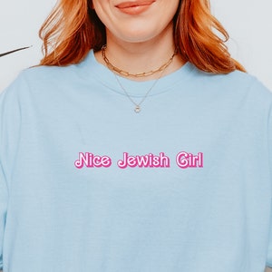 Può includere: T-shirt azzurro con la scritta rosa "Nice Jewish Girl". La maglietta è realizzata in materiale morbido e ha maniche corte. La modella indossa jeans blu e scarpe da ginnastica bianche.