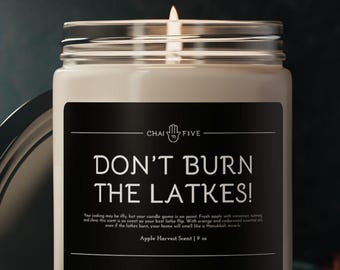 May include: A white candle in a glass jar with a black label that reads "Don't Burn The Latkes!" The candle is lit and the flame is visible. The candle is on a dark surface.
