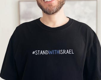 Könnte beinhalten: Schwarzes T-Shirt mit dem Text "#STANDWITHISRAEL" in Blau und Weiß. Das T-Shirt hat einen Rundhalsausschnitt.