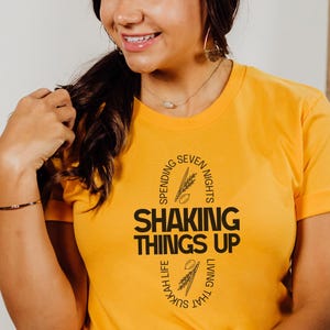 Puede incluir: Camiseta de color amarillo mostaza con el texto negro "SHAKING THINGS UP", rodeado de texto e ilustraciones de trigo. La camiseta es de manga corta y tiene cuello redondo. La camiseta se lleva con vaqueros azul oscuro.