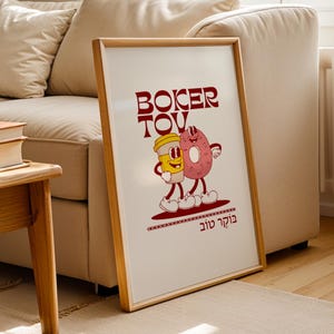 May include: A framed poster featuring a cartoon illustration of a coffee cup and a donut with arms and legs, standing together. The poster has the words "Boker Tov" in red, with Hebrew text below. The frame is light brown.