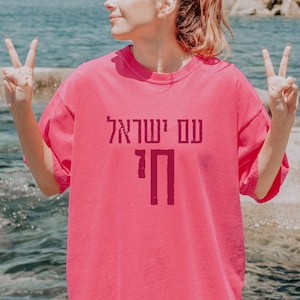 May include: A bright pink t-shirt with Hebrew text. The shirt has a relaxed fit and short sleeves. The text is in a darker shade of pink and is centered on the front of the shirt. The background shows water and rocks.