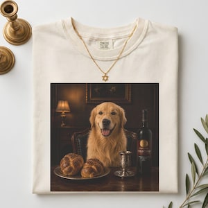 May include: An ivory t-shirt featuring a graphic print of a golden retriever wearing glasses, sitting at a table with two braided breads, a silver goblet, and a wine bottle. A gold-toned Star of David necklace rests on the shirt neckline.
