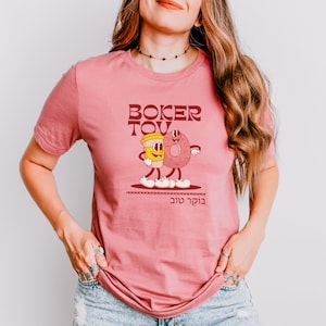 May include: A dusty rose t-shirt featuring a cartoon illustration of a coffee cup and a donut with arms and legs, walking together. The words "Boker Tov" are printed above and below the illustration in a brown font.