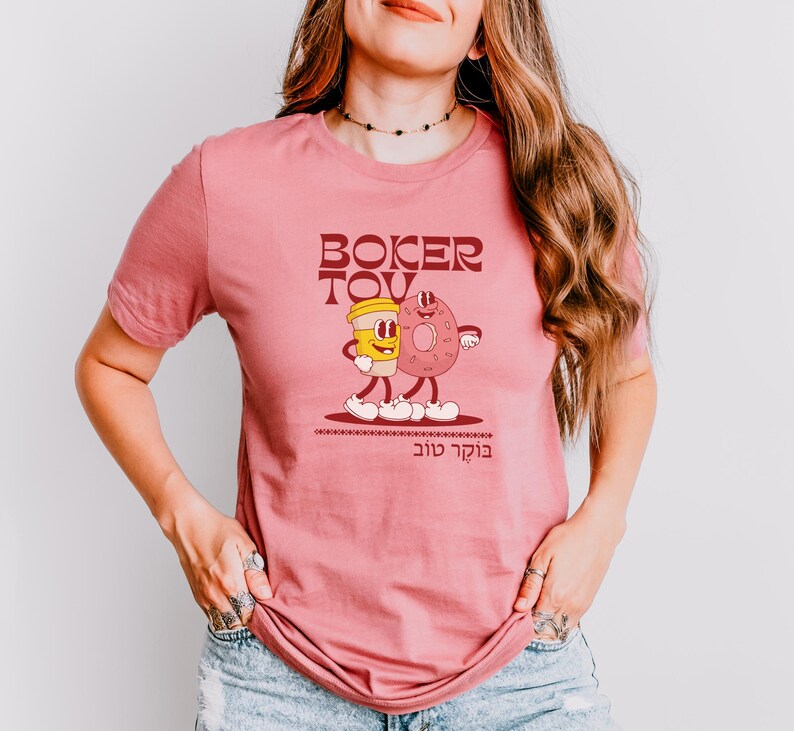 May include: A dusty rose t-shirt featuring a cartoon illustration of a coffee cup and a donut with arms and legs, walking together. The words "Boker Tov" are printed above and below the illustration in a brown font.
