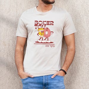 May include: Beige t-shirt featuring a cartoon illustration of a coffee cup and a donut with the text "Boker Tov" in maroon. The design includes the Hebrew phrase "Boker Tov" below the illustration. The shirt is short-sleeved.