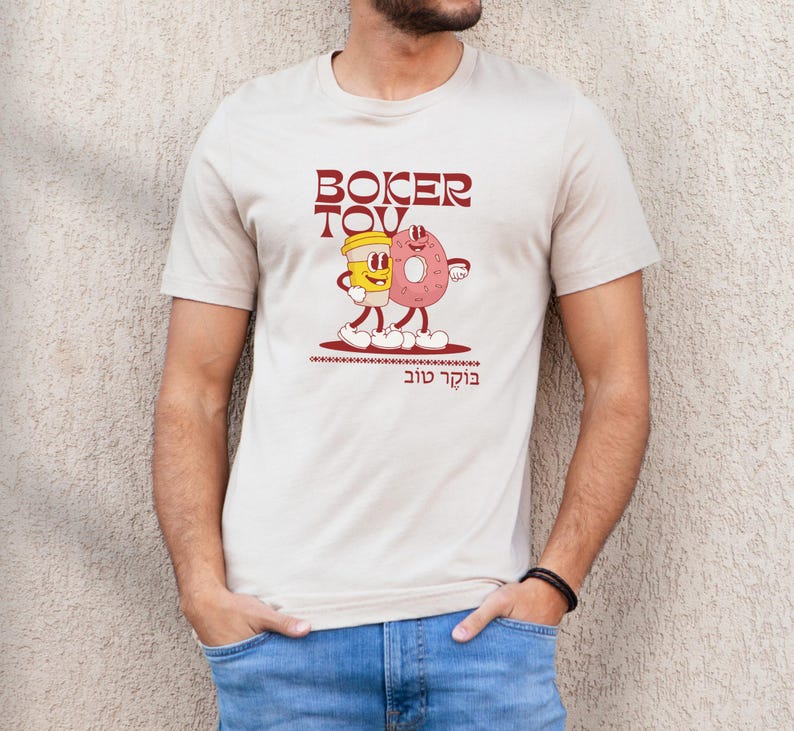 May include: Beige t-shirt featuring a cartoon illustration of a coffee cup and a donut with the text "Boker Tov" in maroon. The design includes the Hebrew phrase "Boker Tov" below the illustration. The shirt is short-sleeved.