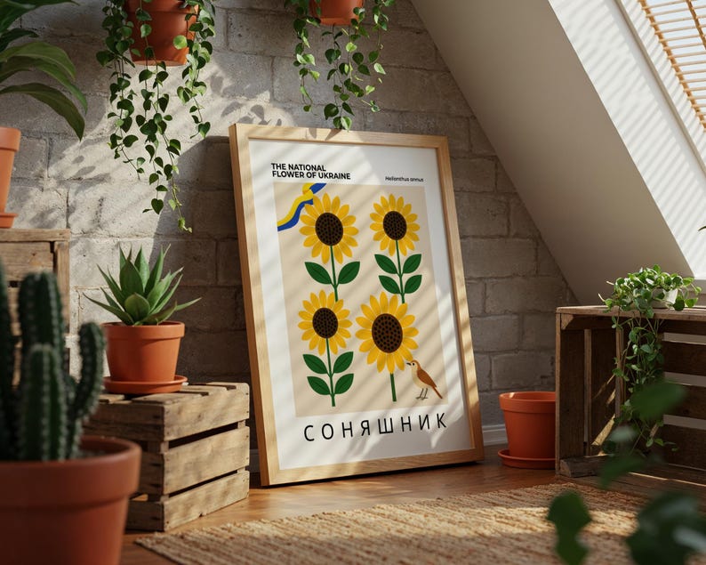 May include: A framed print featuring four stylized sunflowers with green stems and leaves. The text reads "THE NATIONAL FLOWER OF UKRAINE" and "СОНЯШНИК". A small bird is perched near the bottom. The print is displayed in a room with plants and wooden crates.