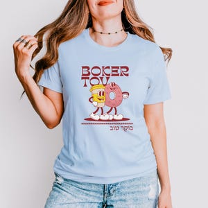 May include: Light blue t-shirt featuring a cartoon illustration of a coffee cup and a donut with arms and legs. The text "BOKER TOV" is above the illustration, and Hebrew text is below. The design is in shades of red, yellow, and pink.