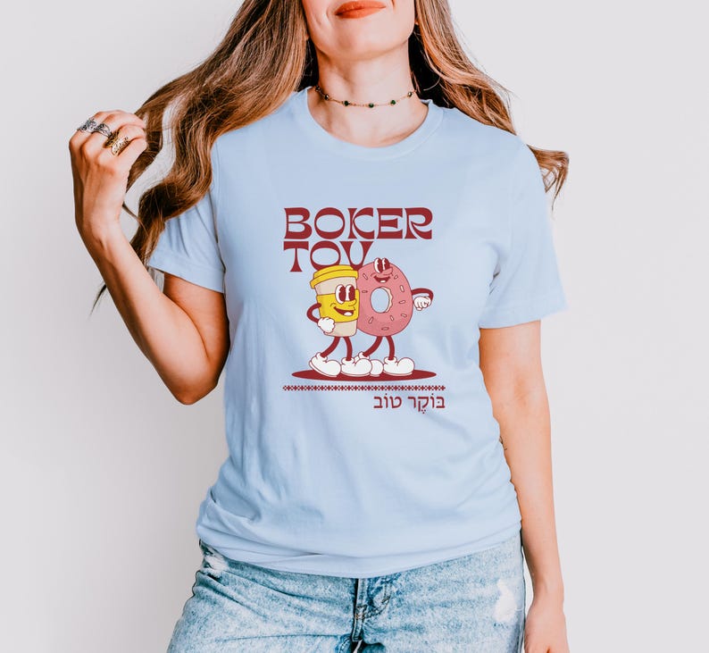 May include: Light blue t-shirt featuring a cartoon illustration of a coffee cup and a donut with arms and legs. The text "BOKER TOV" is above the illustration, and Hebrew text is below. The design is in shades of red, yellow, and pink.