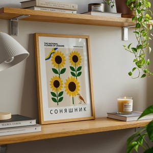 May include: A framed poster featuring sunflowers, the national flower of Ukraine, with text in English and Ukrainian. The poster is displayed on a wooden shelf with books, a lamp, and a lit candle. A globe and hanging plant are also visible.