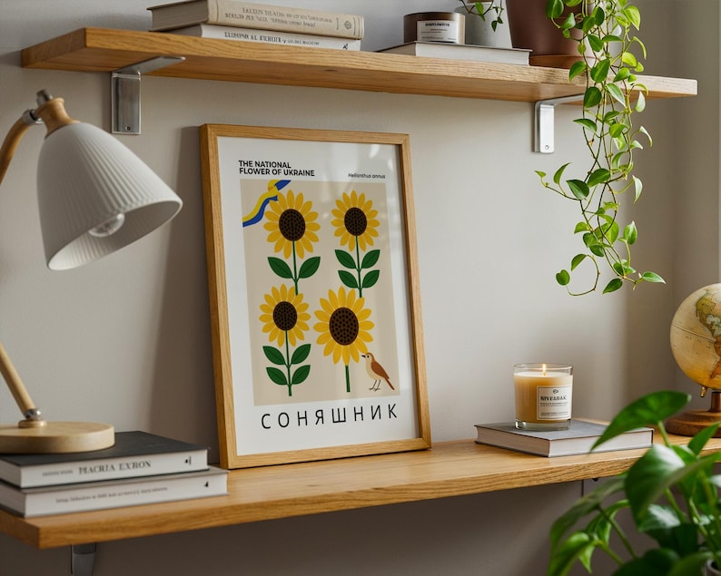 May include: A framed poster featuring sunflowers, the national flower of Ukraine, with text in English and Ukrainian. The poster is displayed on a wooden shelf with books, a lamp, and a lit candle. A globe and hanging plant are also visible.