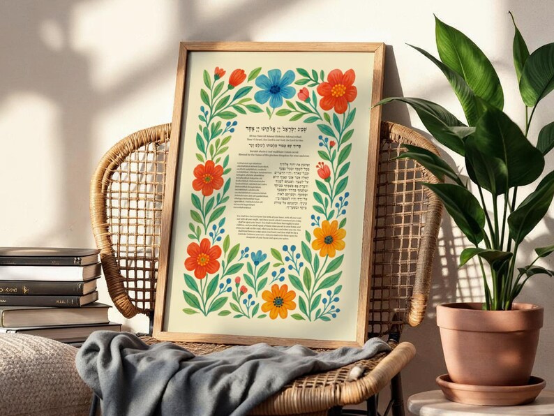 Peut inclure: Une affiche encadr&eacute;e avec des illustrations florales rouges, bleues et jaunes. L'&oelig;uvre est entour&eacute;e d'une bordure florale et comprend du texte en h&eacute;breu. L'affiche est expos&eacute;e sur une chaise en osier &agrave; c&ocirc;t&eacute; d'une plante en pot.