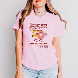 May include: Pink t-shirt with a cartoon graphic of a coffee cup and a donut with the text "Boker Tov" in maroon. The graphic has a retro style. The shirt is made of a soft material.