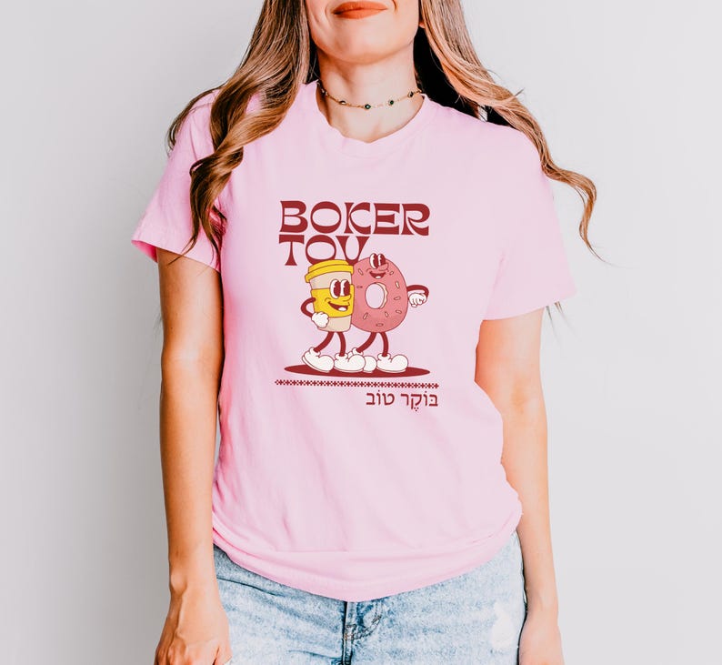 May include: Pink t-shirt with a cartoon graphic of a coffee cup and a donut with the text "Boker Tov" in maroon. The graphic has a retro style. The shirt is made of a soft material.