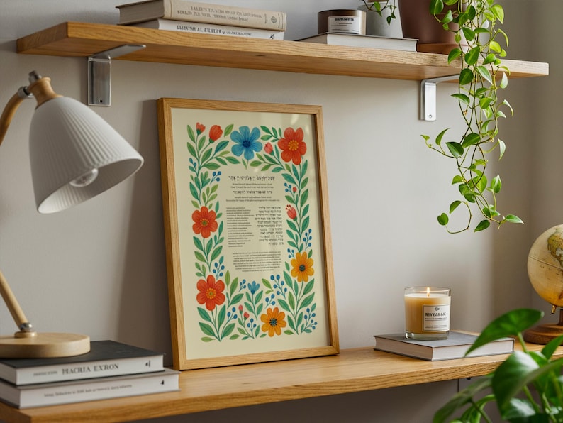 Peut inclure: Une affiche encadr&eacute;e avec une bordure florale dans un cadre en bois. L'affiche pr&eacute;sente du texte en h&eacute;breu et des fleurs color&eacute;es en rouge, bleu et jaune. L'affiche est expos&eacute;e sur une &eacute;tag&egrave;re en bois avec une lampe et des livres.