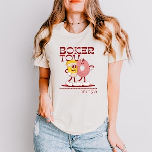 May include: Cream-colored t-shirt featuring a cartoon illustration of a coffee cup and a donut with arms and legs, walking together. The text "BOKER TOV" is above the illustration in maroon, with Hebrew text below. The shirt is worn by a person.