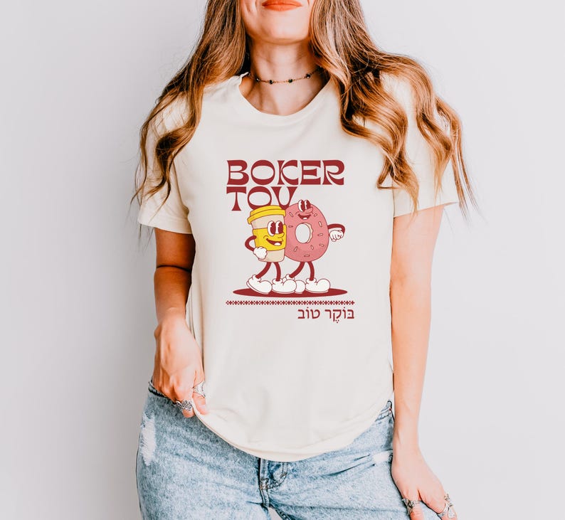 May include: Cream-colored t-shirt featuring a cartoon illustration of a coffee cup and a donut with arms and legs, walking together. The text "BOKER TOV" is above the illustration in maroon, with Hebrew text below. The shirt is worn by a person.