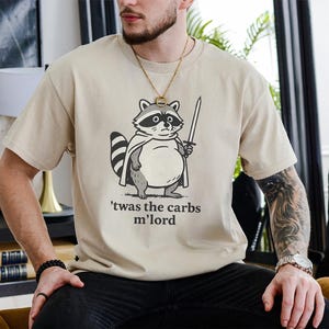 May include: A beige t-shirt with a cartoon raccoon holding a sword and wearing a cape. The text on the shirt reads "'twas the carbs m'lord". The raccoon is in black and white.