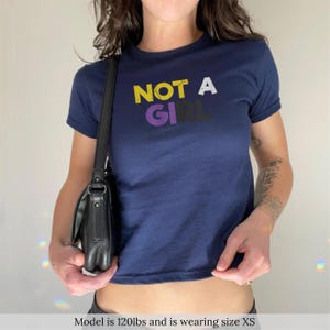 Op de afbeelding: Marineblauwe crop top met de tekst "NOT A GIRL" in geel, paars en zwart. De top heeft korte mouwen en een ronde hals. Een zwarte schoudertas hangt aan de linkerkant.