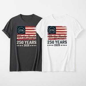 Puede incluir: Dos camisetas, una gris oscuro y otra blanca, cada una con un diseño patriótico. El diseño incluye una bandera estadounidense desgastada, el texto "1776", "We the People", "250 YEARS" y "2026" con estrellas. Un diseño para ropa patriótica.
