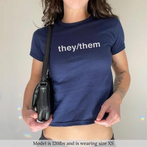 Könnte beinhalten: Ein marineblaues Crop-Top mit dem weißen Text "they/them". Das Shirt hat kurze Ärmel und einen Rundhalsausschnitt. Eine schwarze Umhängetasche wird getragen. Das Model trägt Größe XS.