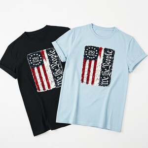 May include: Two t-shirts, one black and one light blue, each with a distressed American flag design. The flag graphic includes the text "250 Years" and "We the People". The shirts are short-sleeved with a crew neck.